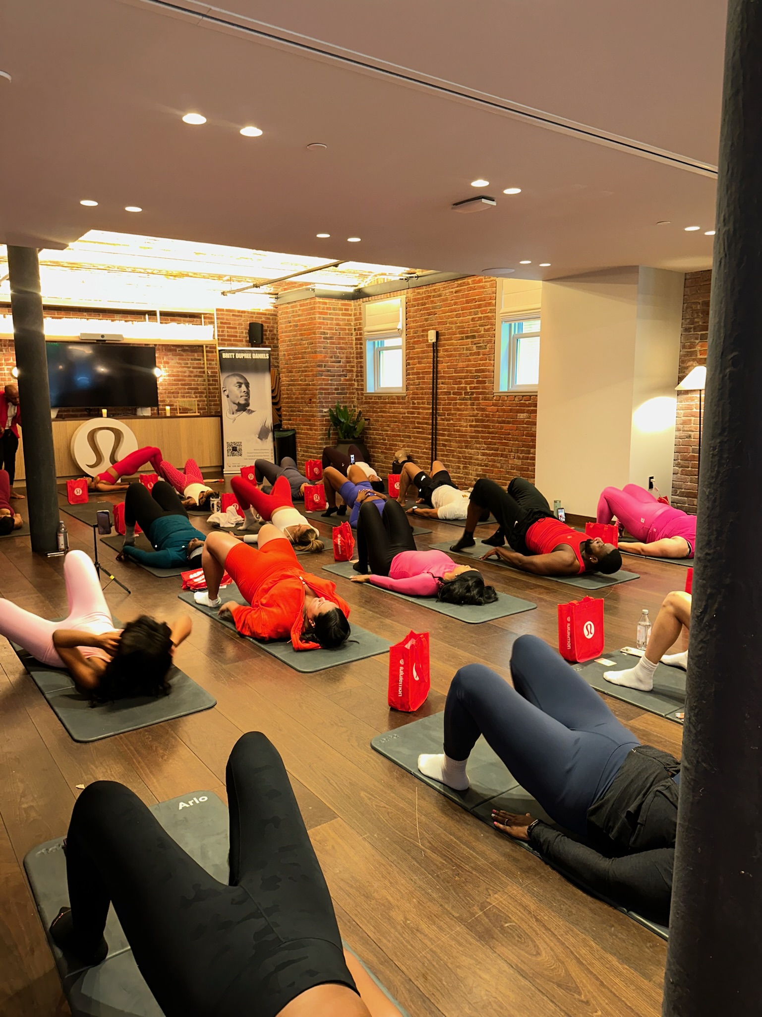 A group of people in athletic wear are lying on yoga mats doing fitness exercises in a studio with brick walls and natural light. Red Lululemon gift bags are next to each person.