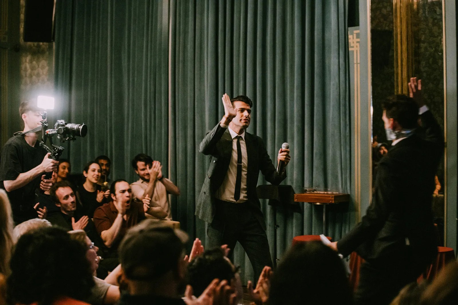 A man in a suit holds a microphone and gives a high five to another man on stage in front of an applauding audience, with a cameraman filming the event.