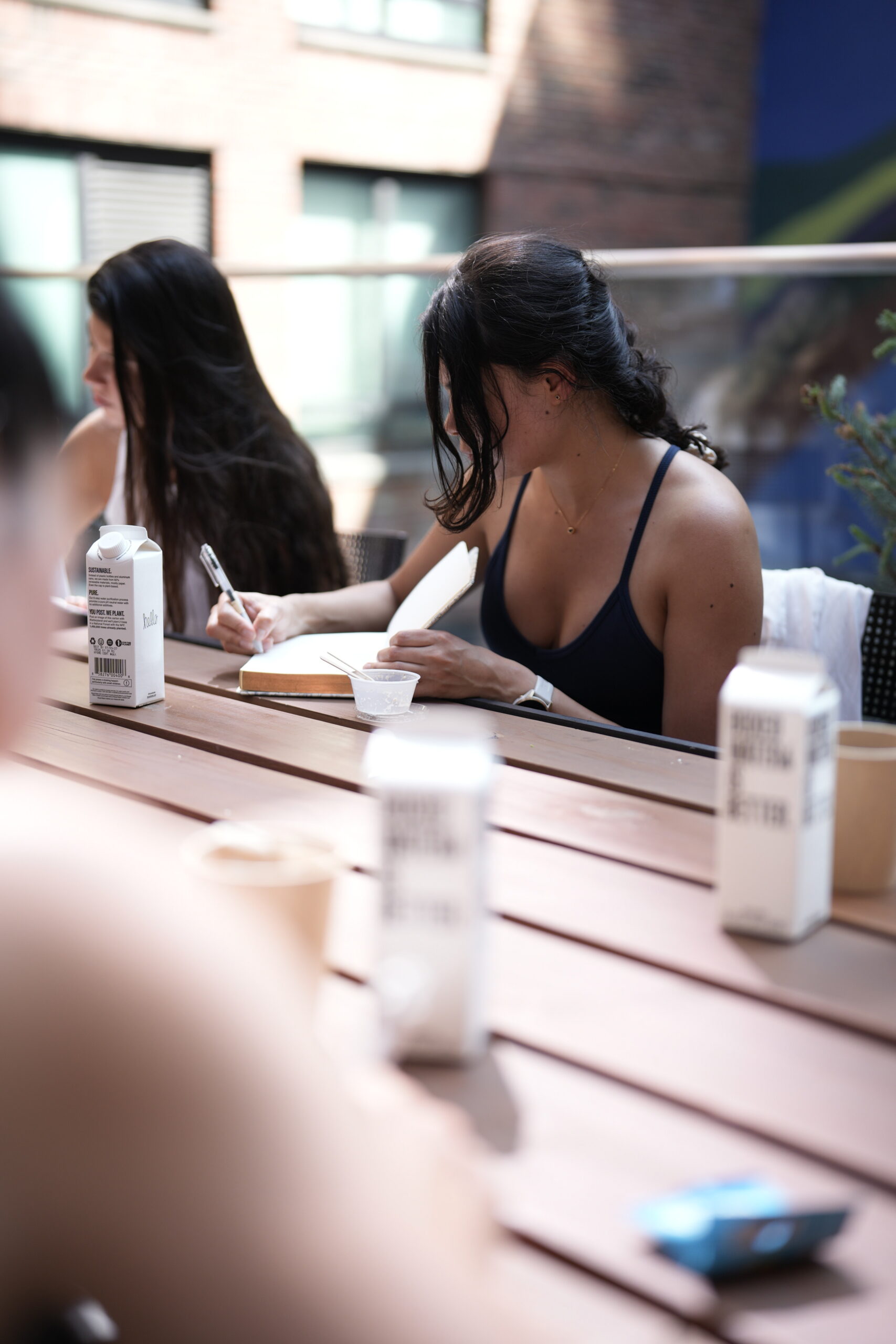 A woman sits at an outdoor table, writing in a notebook with cartons of boxed water and a yogurt cup in front of her.