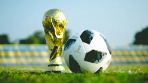 A gold FIFA World Cup trophy stands next to an Adidas Telstar 18 soccer ball on a grassy field, with blurred stadium seats in the background.