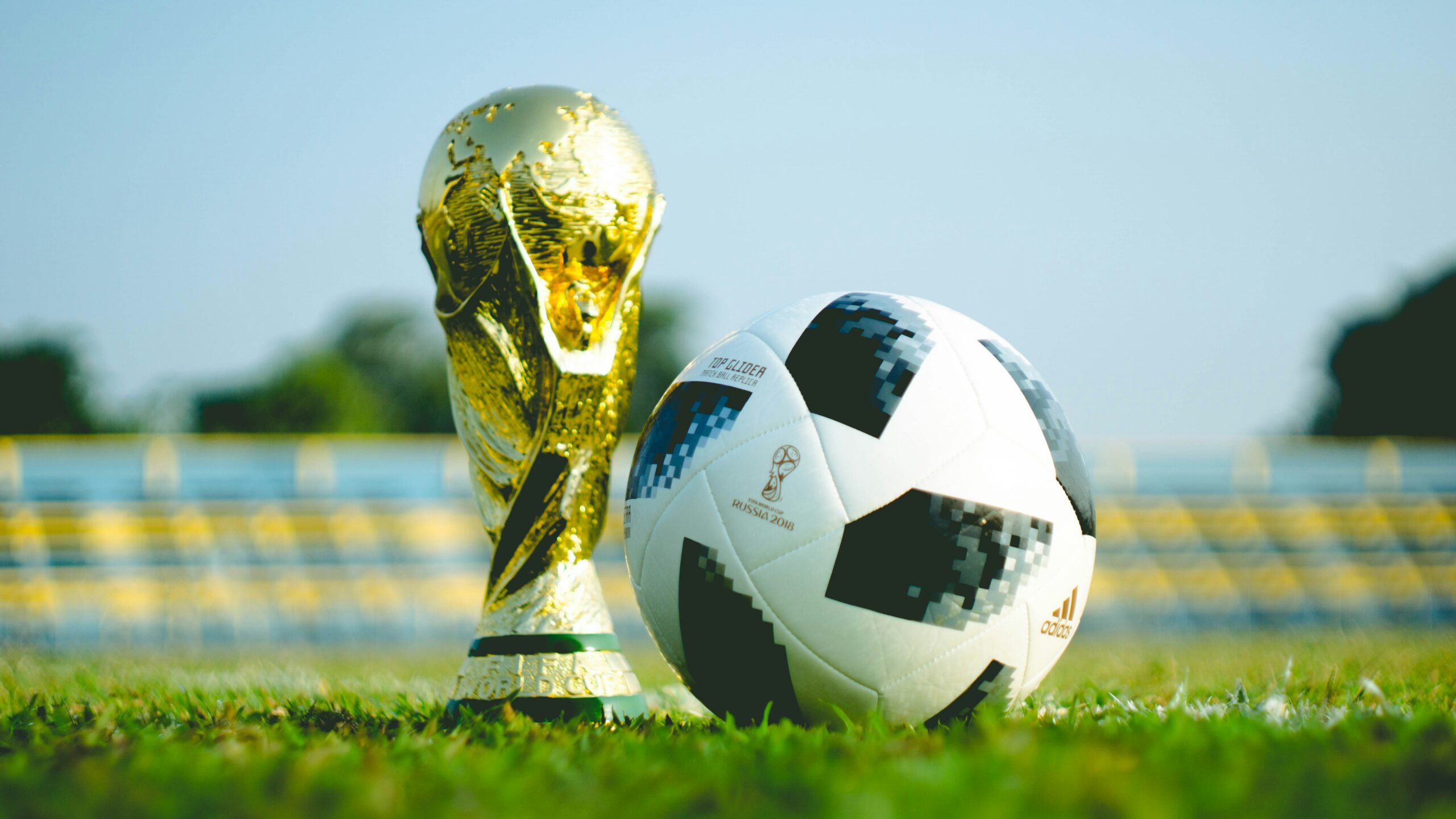 A gold FIFA World Cup trophy stands next to an Adidas Telstar 18 soccer ball on a grassy field, with blurred stadium seats in the background.