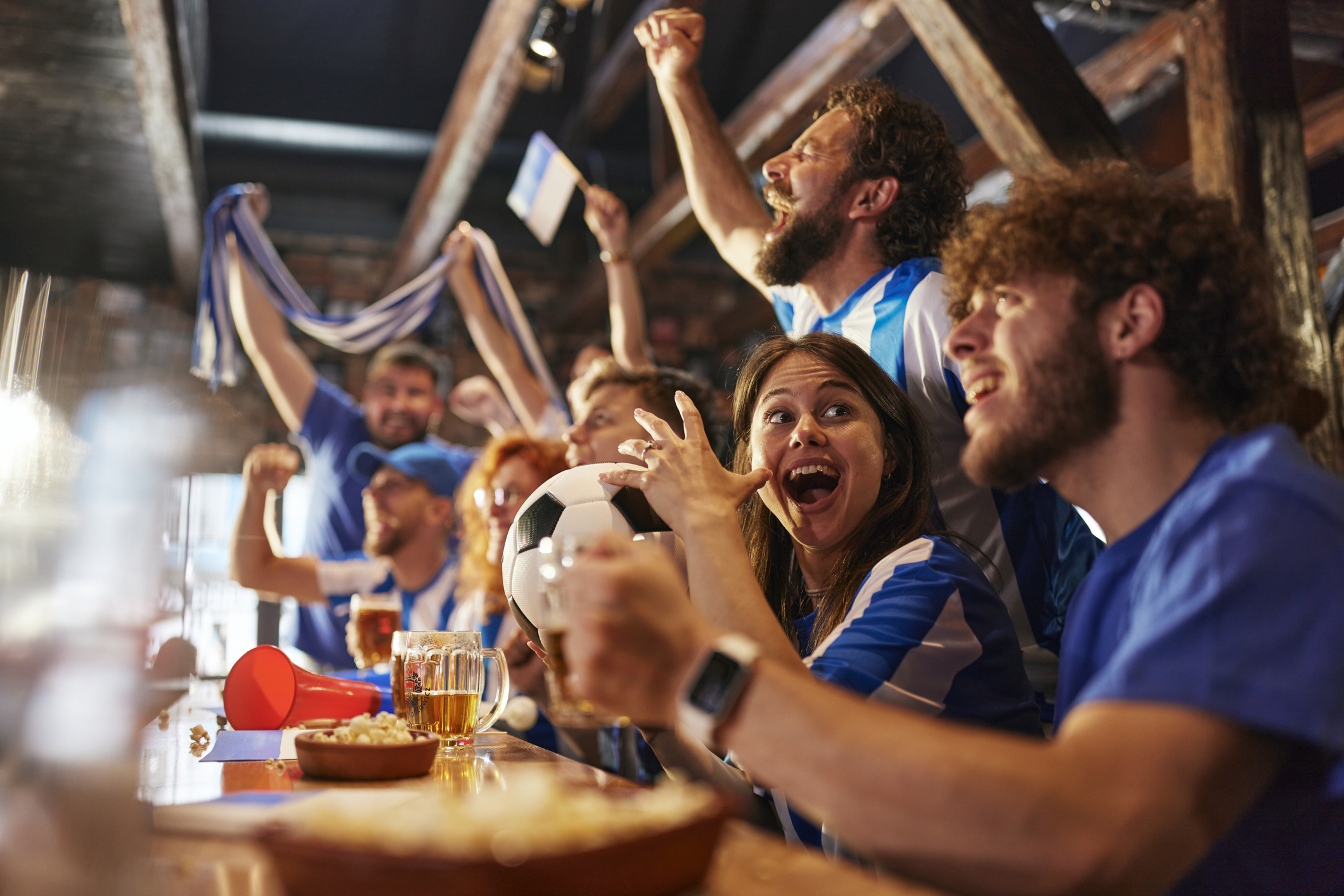 A group of people in blue jerseys sit at a bar, cheering and holding a soccer ball, with drinks and snacks on the table, watching a game.