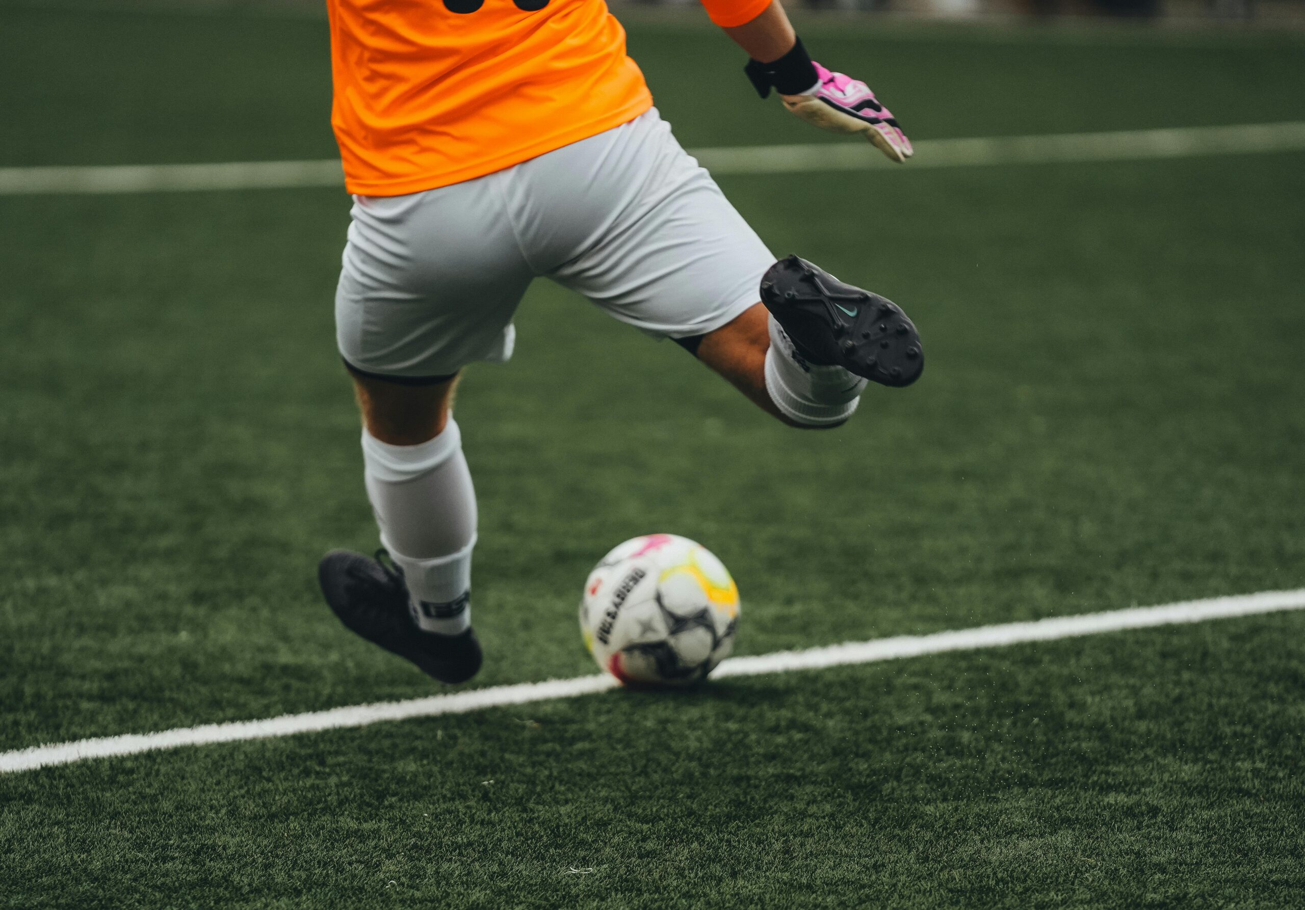 A soccer player in a bright orange jersey prepares to kick a soccer ball on a green field. Only the player's lower body and the ball are visible.