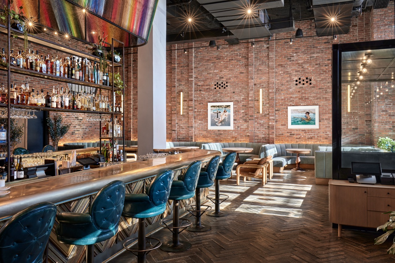 Modern bar and lounge with teal barstools, exposed brick walls, a long counter with liquor bottles, wall art, and natural light streaming through large windows.