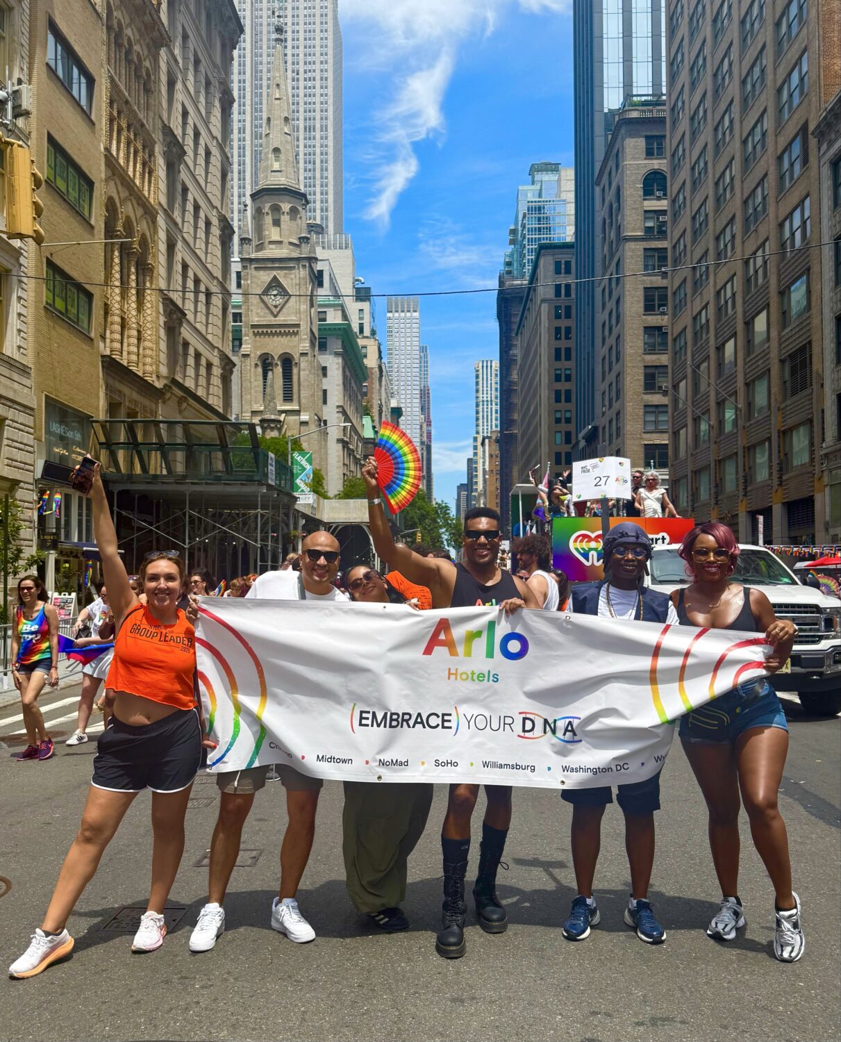 A group of people hold an "Embrace Your DNA" banner for Arlo Hotels on a city street during a parade, with buildings and a parade float visible in the background.