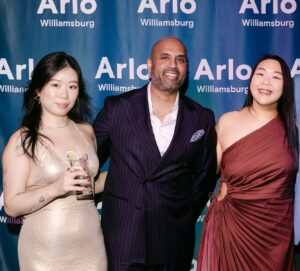 Three people pose together at an event in front of a blue step-and-repeat backdrop with "Arlo Williamsburg" written on it.