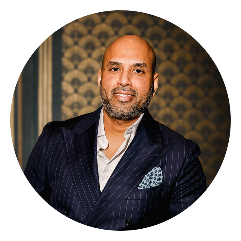 A man with a trimmed beard and bald head wearing a dark pinstripe suit, light shirt, and patterned pocket square, standing in front of a patterned background.