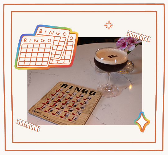 A Bingo card and a cocktail glass with foam art are placed on a table, with a small flower arrangement in the background.