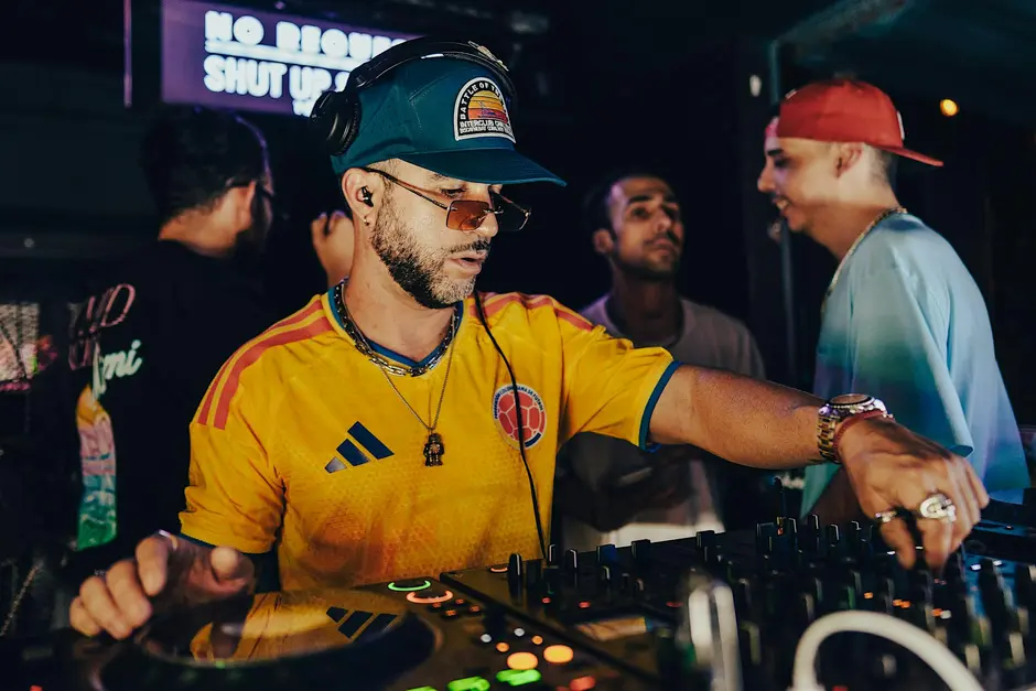 A DJ wearing headphones and a yellow jersey mixes music on a DJ controller, with three people standing and talking in the background at a dimly lit venue.