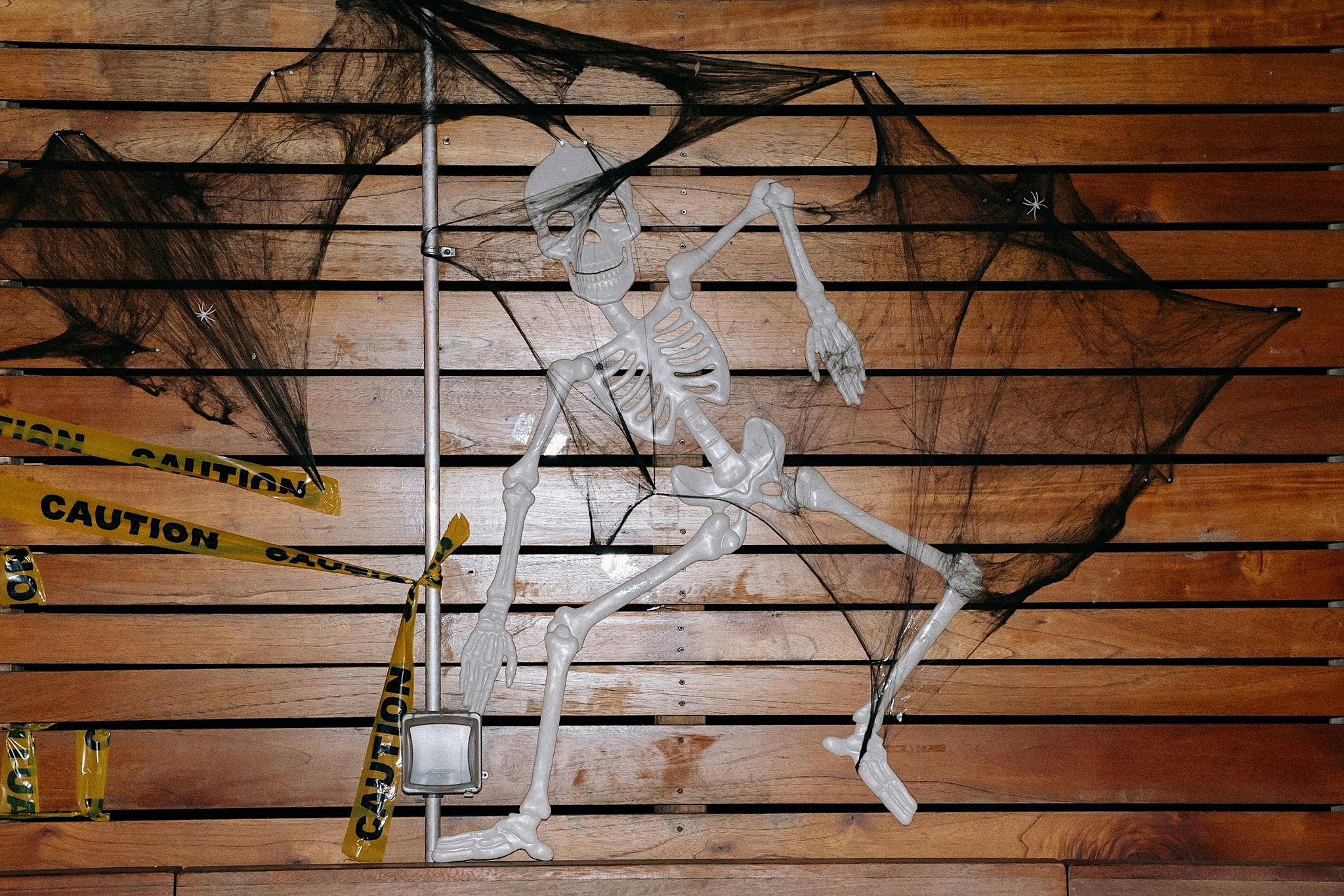 A plastic skeleton and black decorative webbing attached to a slatted wood wall with “CAUTION” tape.