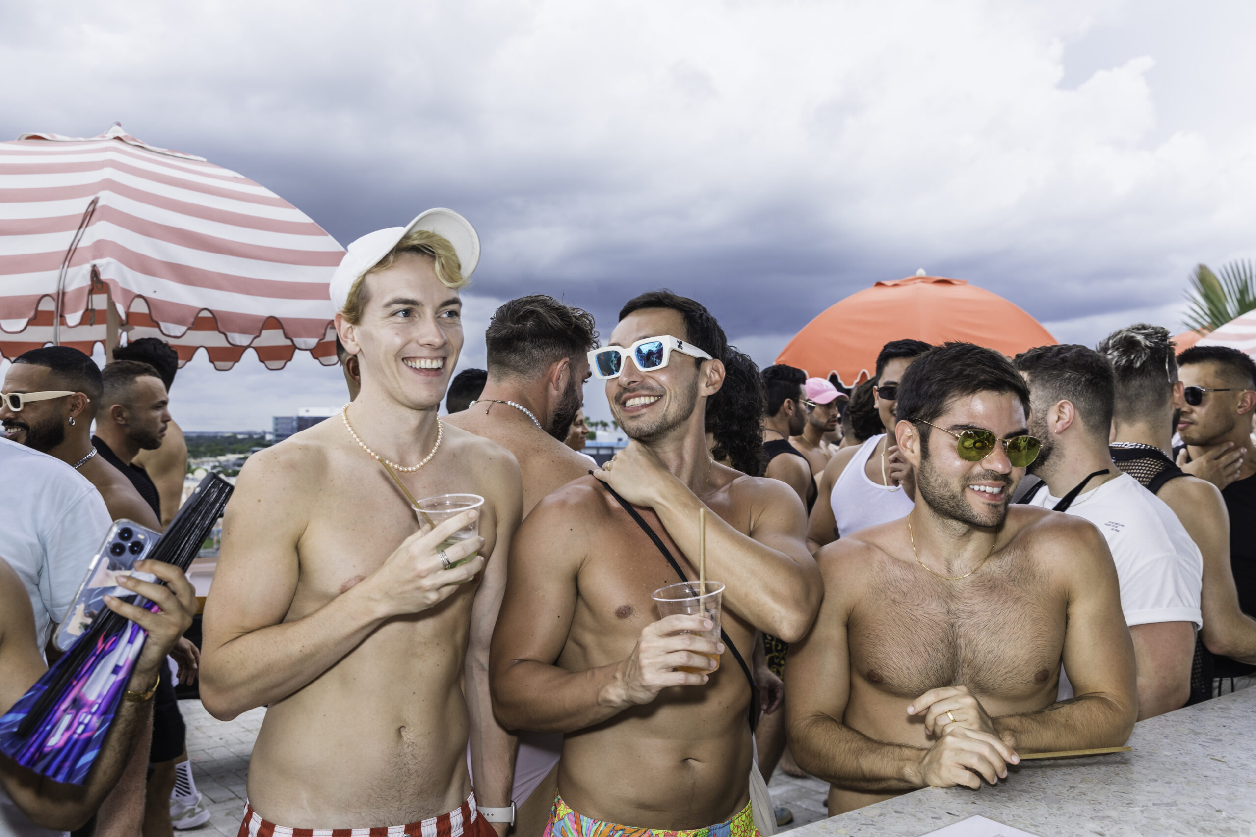 A group of shirtless men enjoying drinks and smiling at an outdoor event with striped umbrellas in the background.