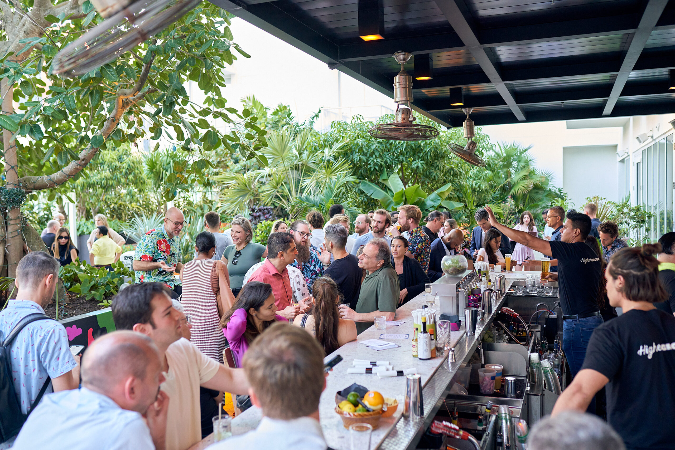 A crowded outdoor bar scene with people socializing and bartenders working behind the counter, surrounded by lush greenery.