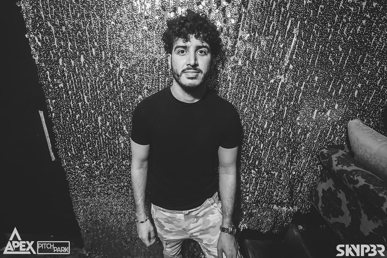 A man with curly hair, wearing a black t-shirt and camouflage pants, stands in front of a glittery backdrop in a dimly lit room. Logos "APEX" and "SKY3R" appear in the corners.