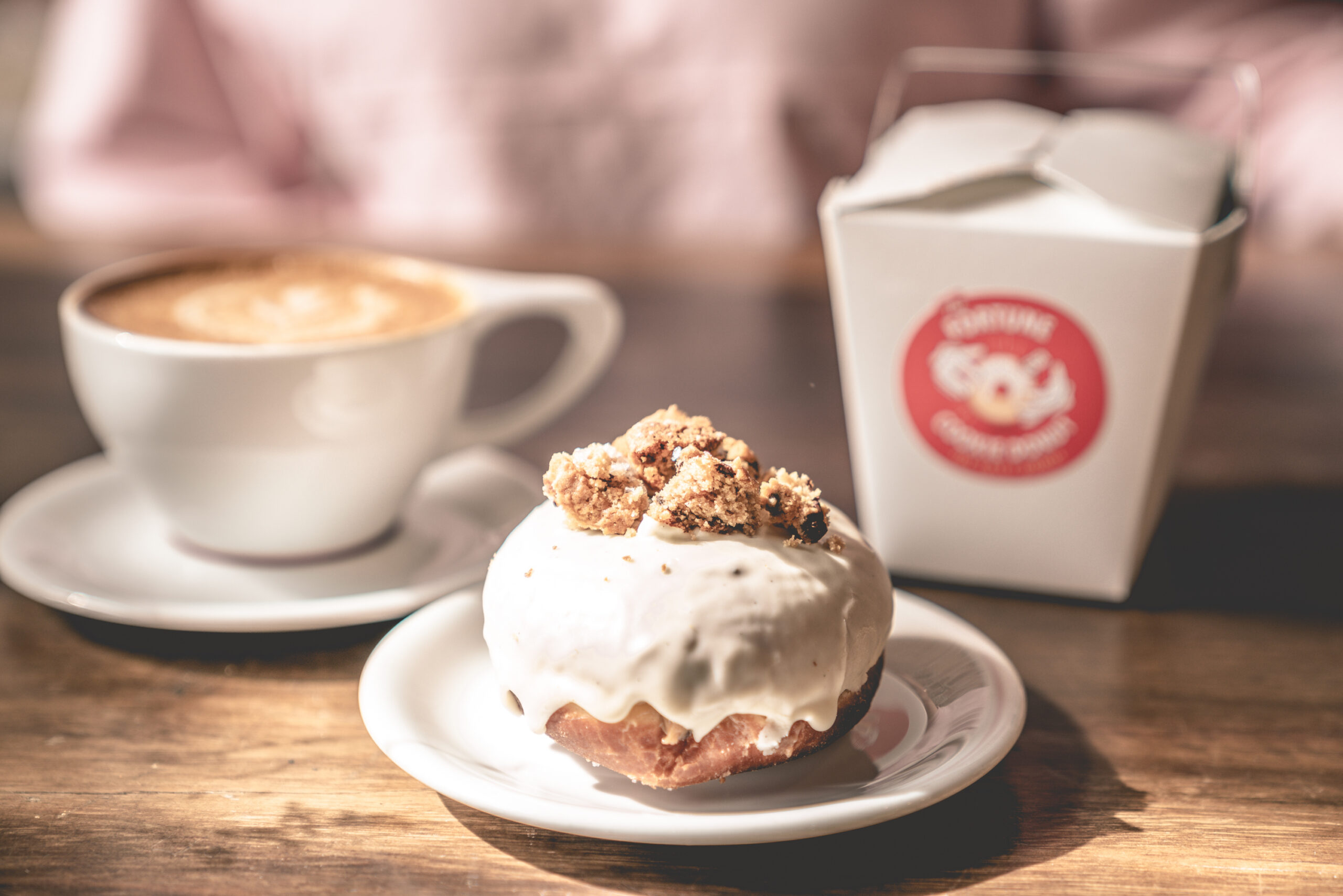 A close-up of a white-glazed specialty donut topped with cookie pieces, served with a latte and a branded takeout box from The Salty Donut.