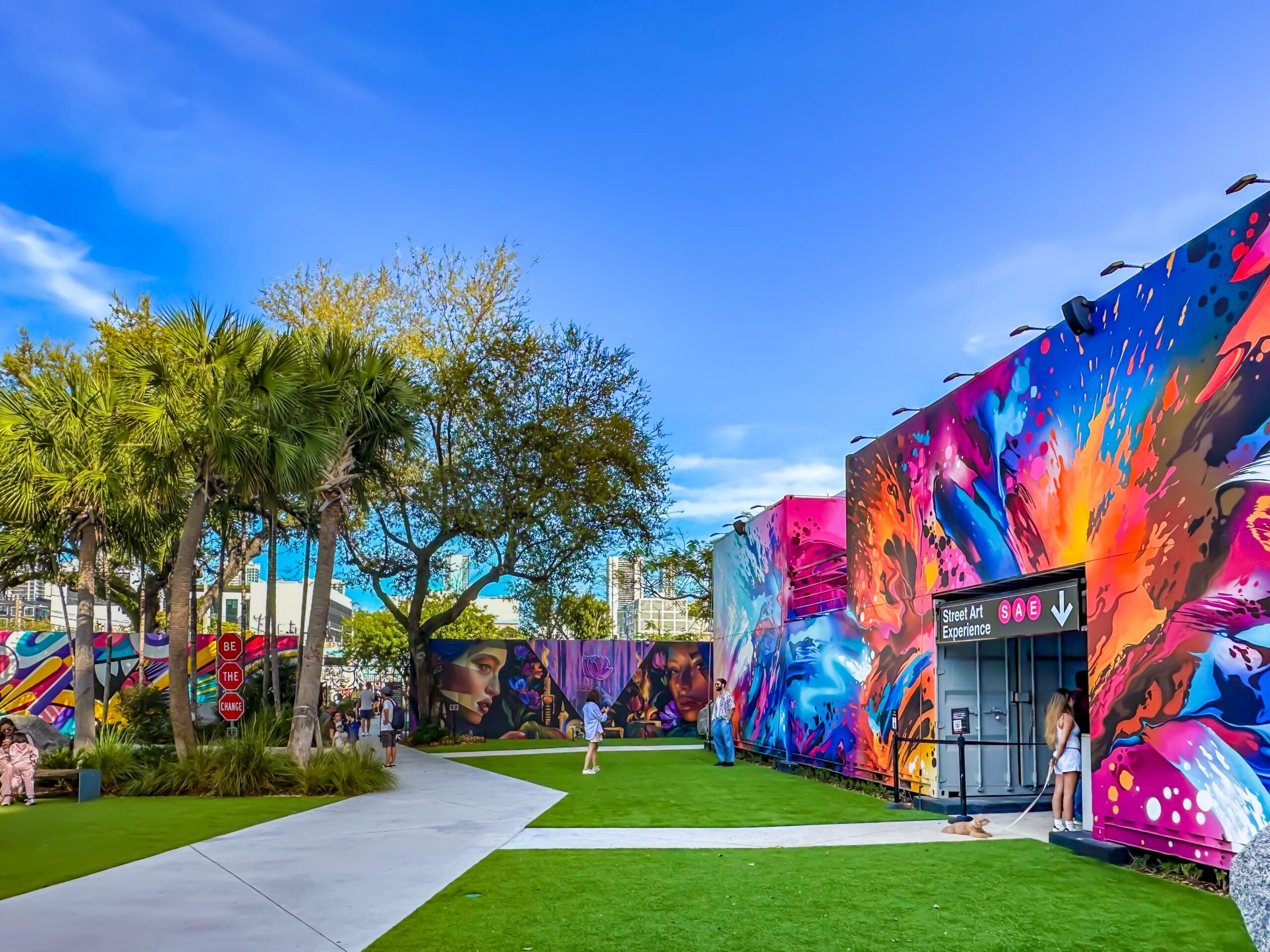 A bright outdoor scene at Wynwood Walls in Miami, showing vibrant, colorful street art murals on warehouse walls surrounding a green lawn with palm trees and a "Street Art Experience" entrance.