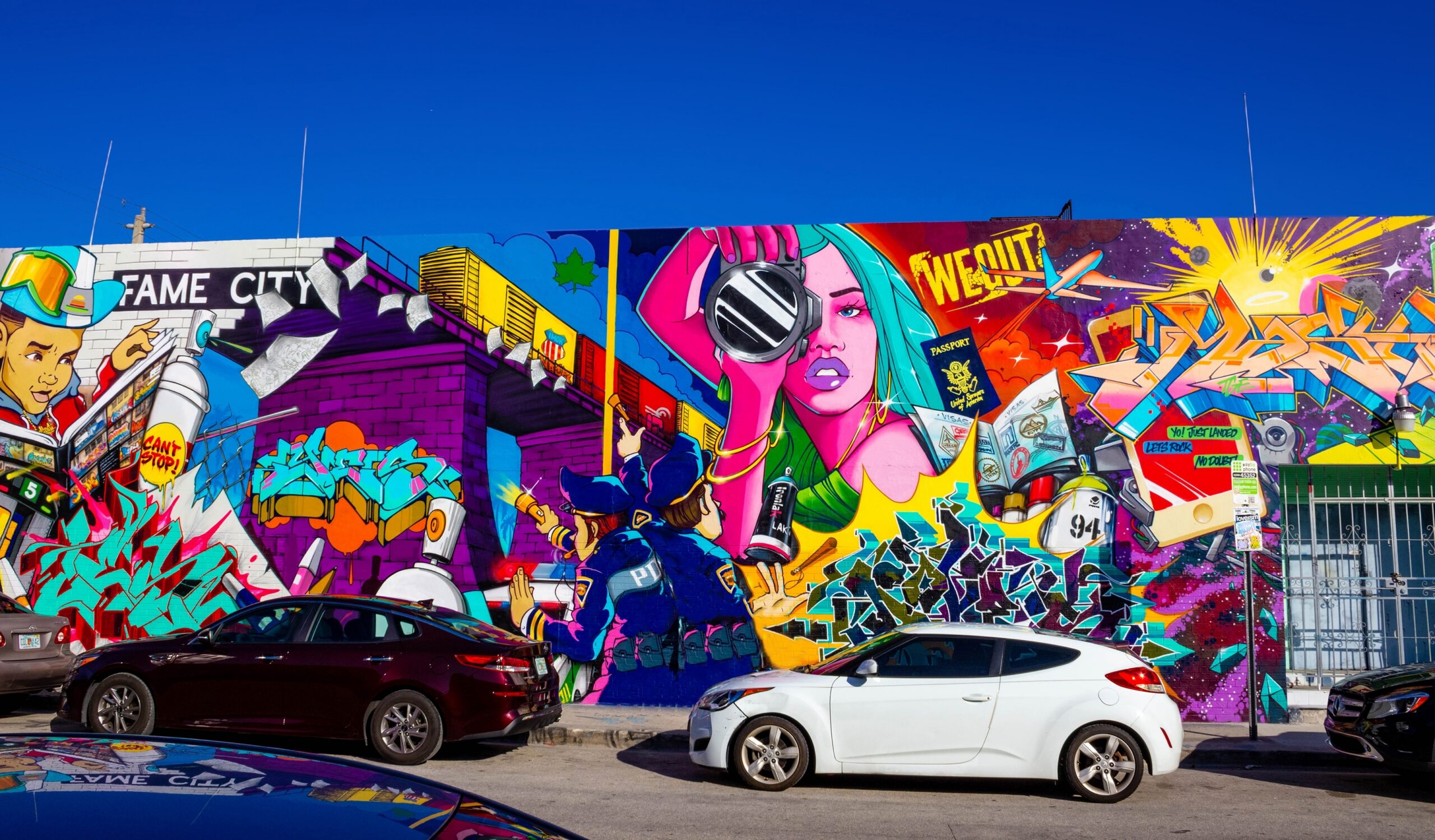 A vibrant, large-scale street art mural in the Wynwood neighborhood, featuring a woman with teal hair holding a camera lens, police officers in uniform, and bold graffiti lettering. The artwork uses a bright palette of purple, pink, and yellow, with several cars parked on the street in the foreground under a clear blue sky.