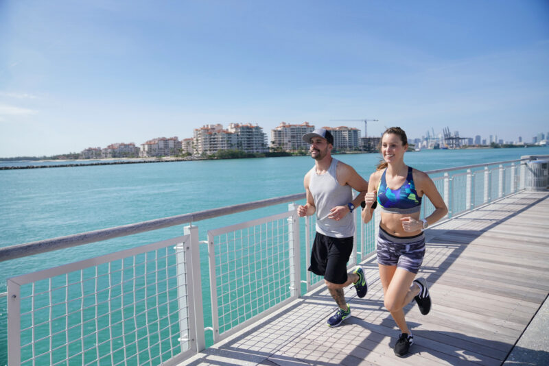 Two people jogging along a sunny waterfront boardwalk with turquoise water and modern buildings in the background, capturing an energetic outdoor fitness scene by the breeze-filled coast.