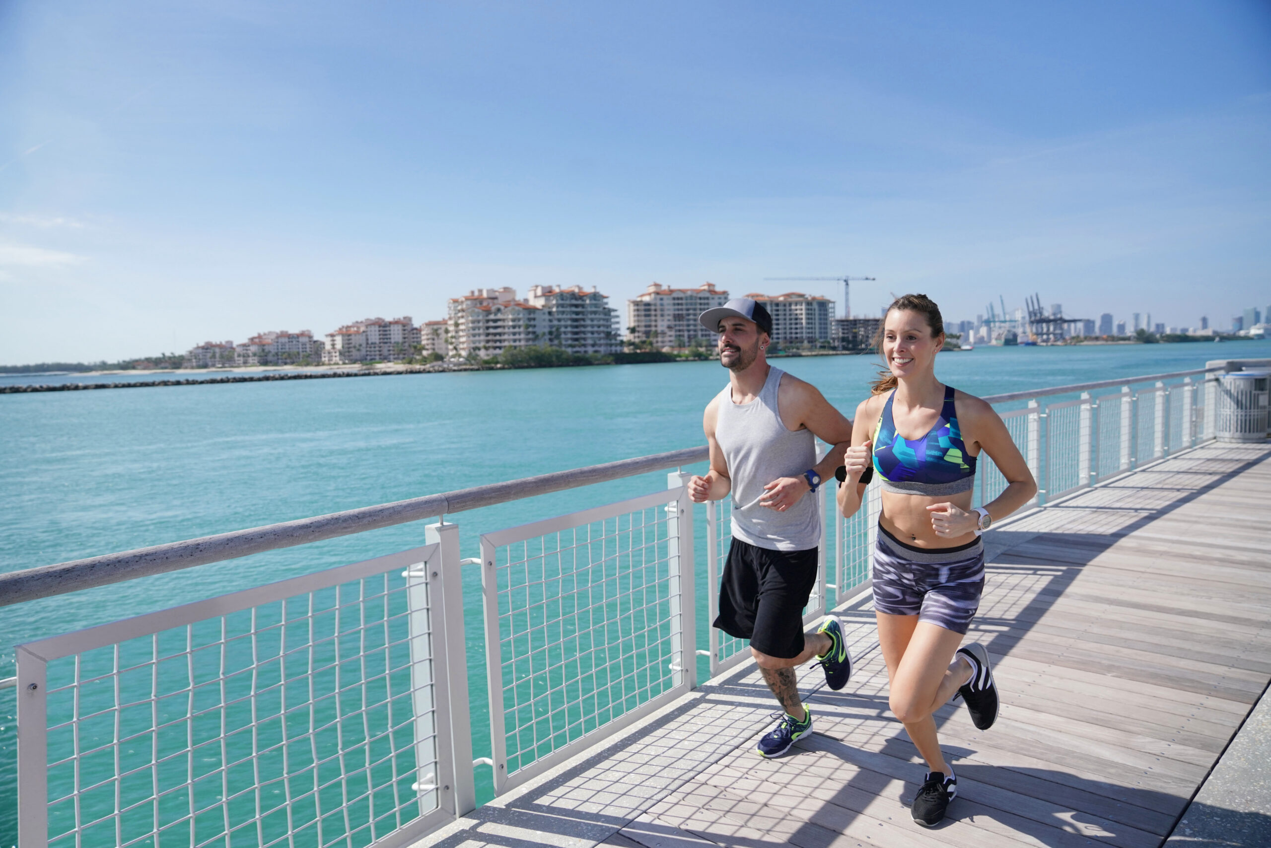 Two people jogging along a sunny waterfront boardwalk with turquoise water and modern buildings in the background, capturing an energetic outdoor fitness scene by the breeze-filled coast.
