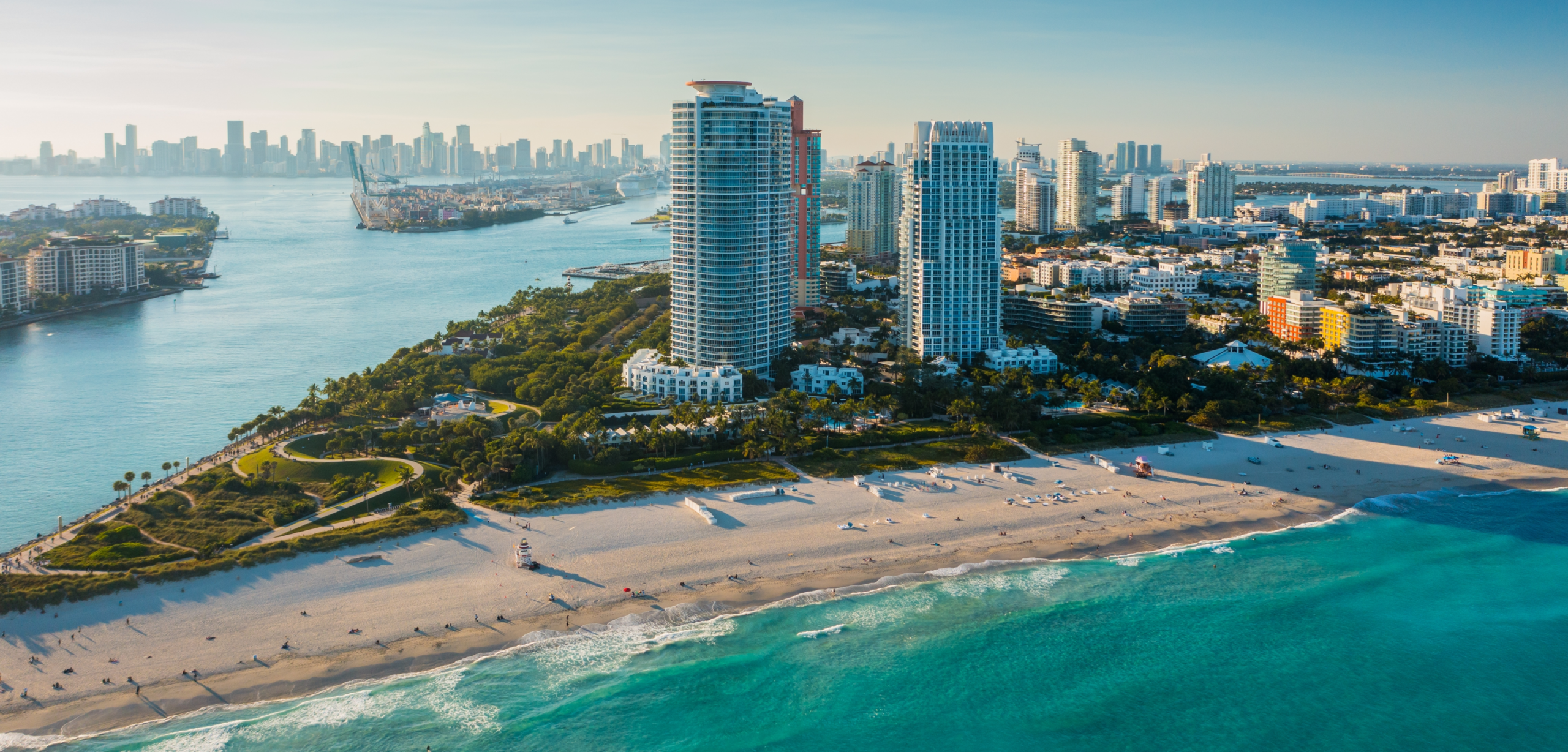 Aerial view of Miami South Beach with turquoise ocean waves, white sandy beaches, waterfront high-rise buildings, and the downtown Miami skyline stretching across the horizon on a clear day.