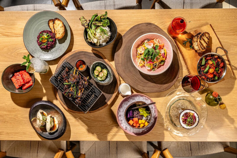 Overhead view of a restaurant table filled with colorful dishes, grilled meats and vegetables, pasta, seafood, desserts, and cocktails.
