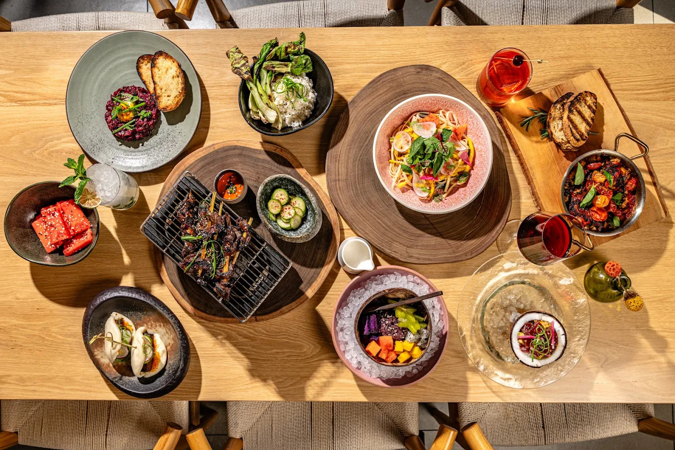 Overhead view of a restaurant table filled with colorful dishes, grilled meats and vegetables, pasta, seafood, desserts, and cocktails.
