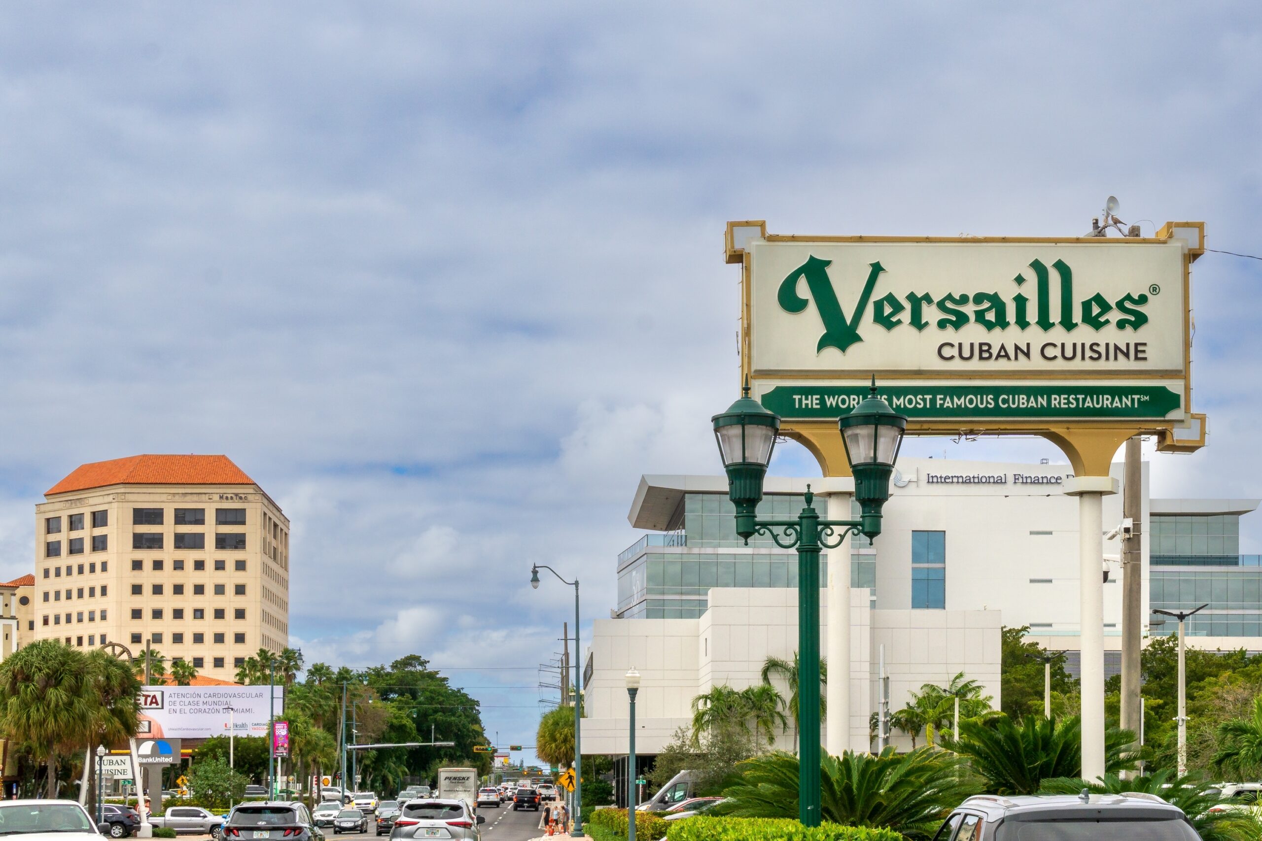 Sign for Versailles Cuban Cuisine restaurant, reading 'The World's Most Famous Cuban Restaurant,' mounted above a street lined with palm trees, traffic, and office buildings in Miami."